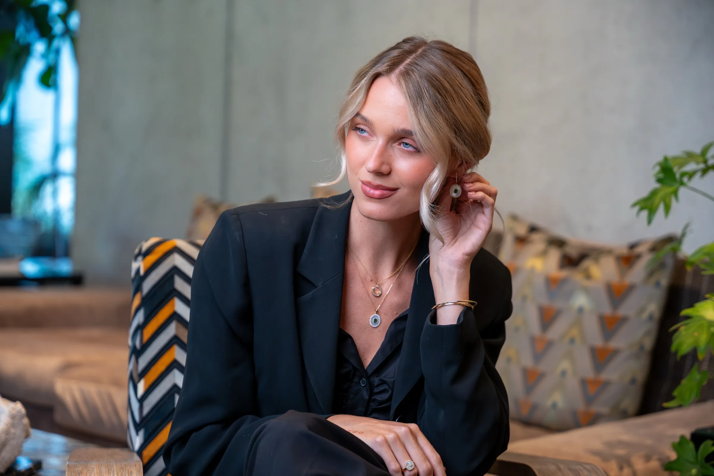 Model in a black blazer wearing layered gold pendant necklaces, earrings and a ring in an Austin hotel lounge